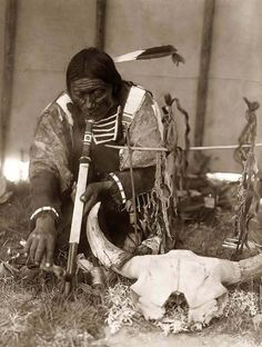 man in traditional dress sitting on the grass smoking a long pipe, tabacco? drying on a stick suspeneded by two other sticks, Buffalo skull, fabric wall and supporting poles in the background.