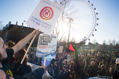 a crowd of demonstrators with an iconic ferris wheel in the background and signs reading, Our World, Our Future, CLIMATE CHANGE IT'S TIME TO ACT, and CAPITALISM CREATES CLIMATE CHAOS