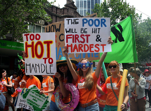 a protest march featuring smiling women in matching orange tshirts, festive swimming pool accessories, and signs that read, TOO HOT TO HANDLE, and, THE POOR WILL BE HIT FIRST & HARDEST